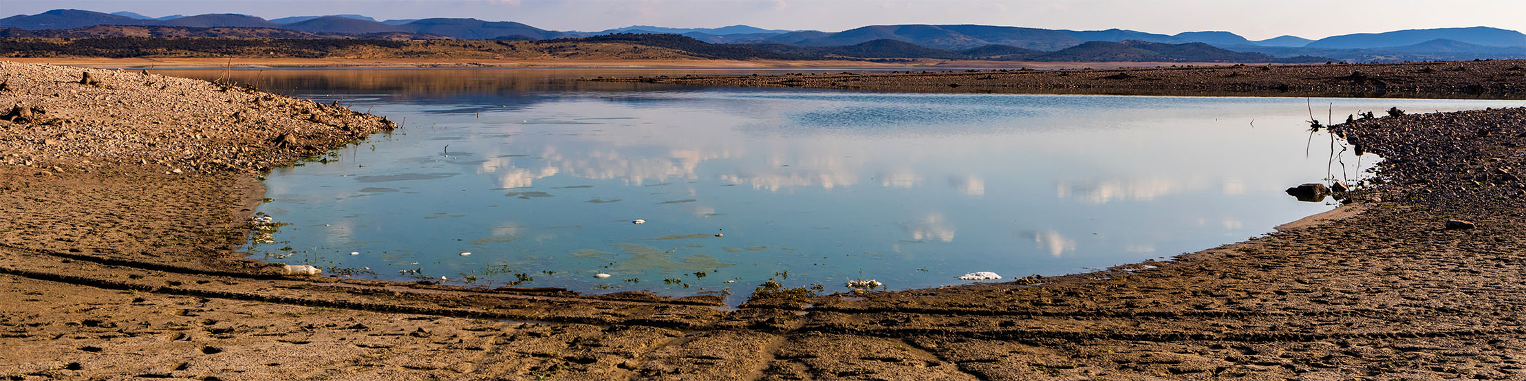 A western reservoir with low water levels and dry, cracked land surrounding with mountains in the background | Adobe Stock